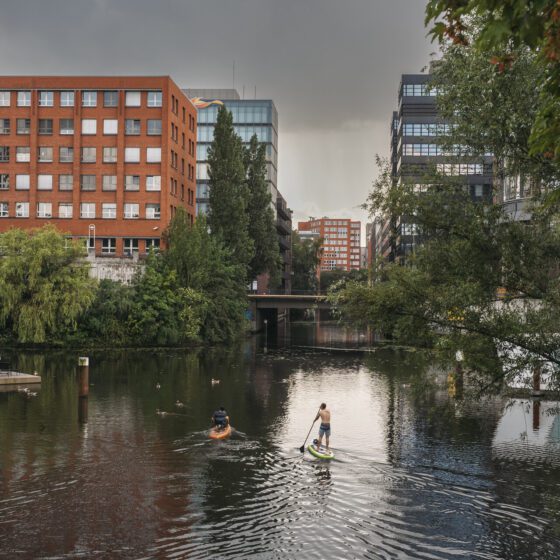 Zwei Personen, die auf einem Stadtfluss mit Stehpaddeln, während der Himmel bewölkt ist und städtische Gebäude im Hintergrund sichtbar sind.