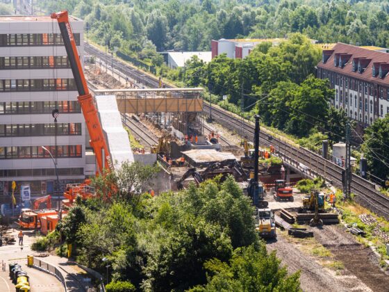 Baustelle mit Kränen neben den Bahngleisen, in der Nähe von Gebäuden und Bäumen.