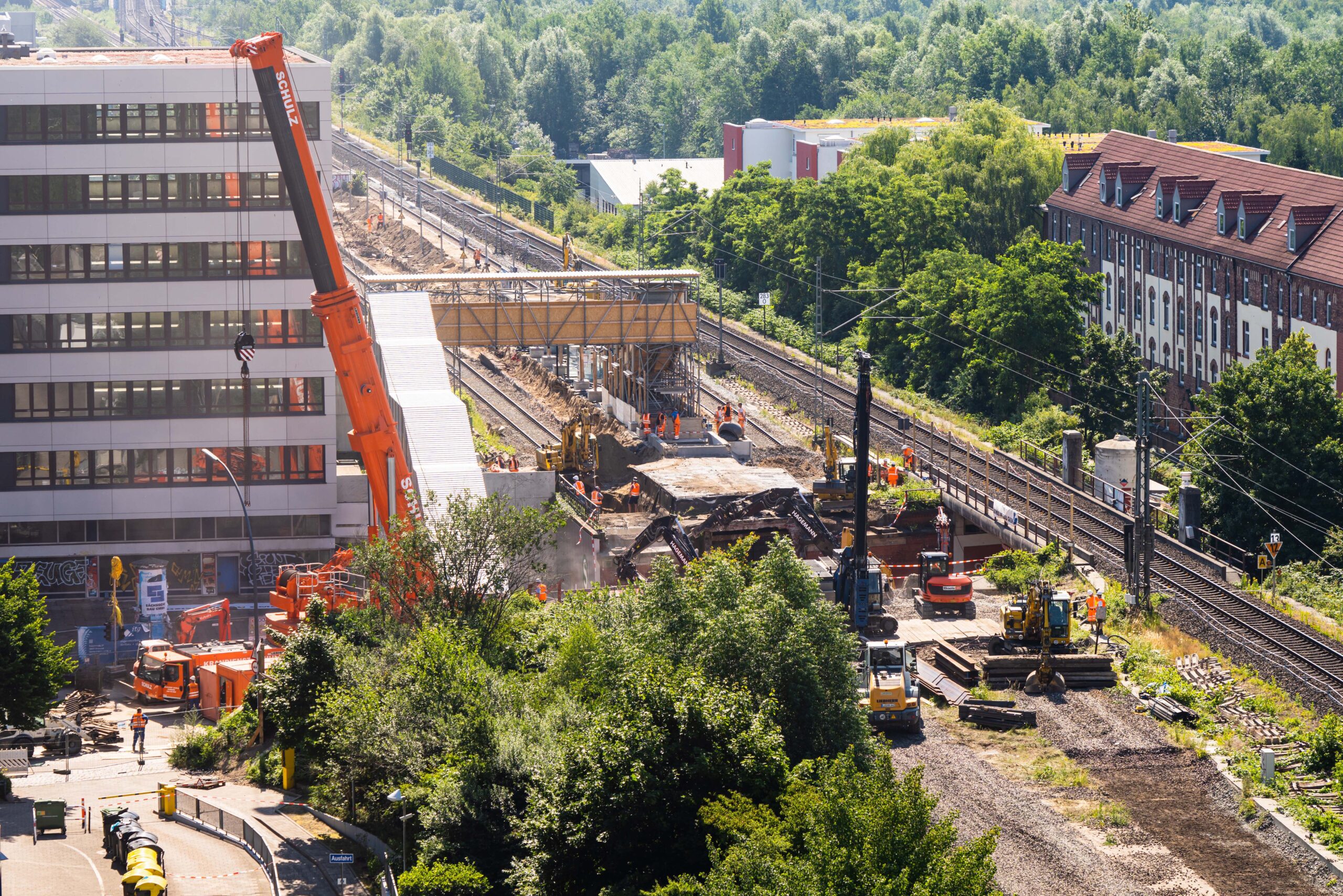 Baustelle mit Kränen neben den Bahngleisen, in der Nähe von Gebäuden und Bäumen.