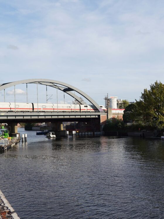 Ein Zug, der eine gewölbte Brücke über einen Fluss überquert, umgeben von industriellen Anlagen.