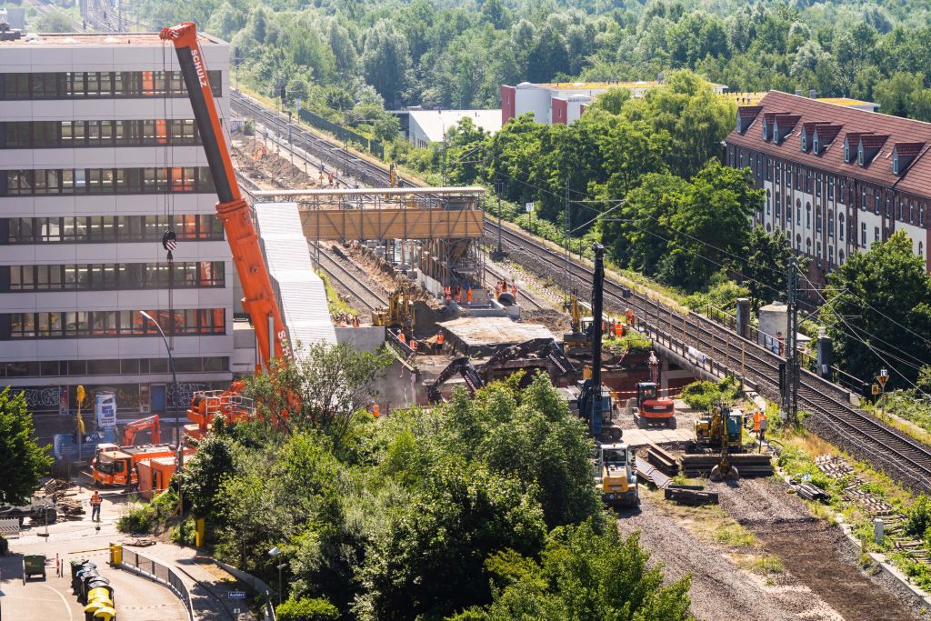 Baustelle mit Kränen an Bahngleisen, nahe bei Gebäuden und Bäumen.